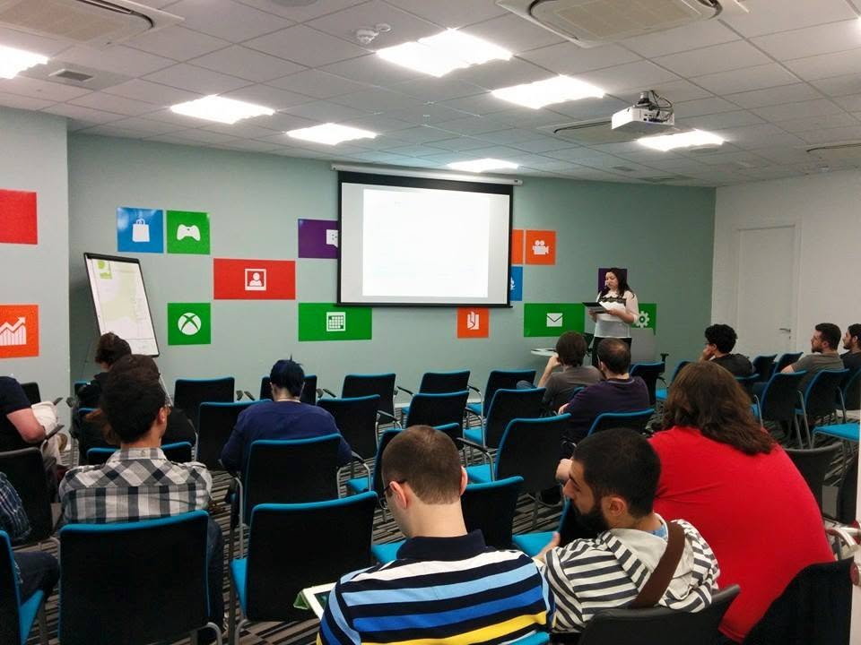 A presenter speaking to a seated audience in a room with a projection screen